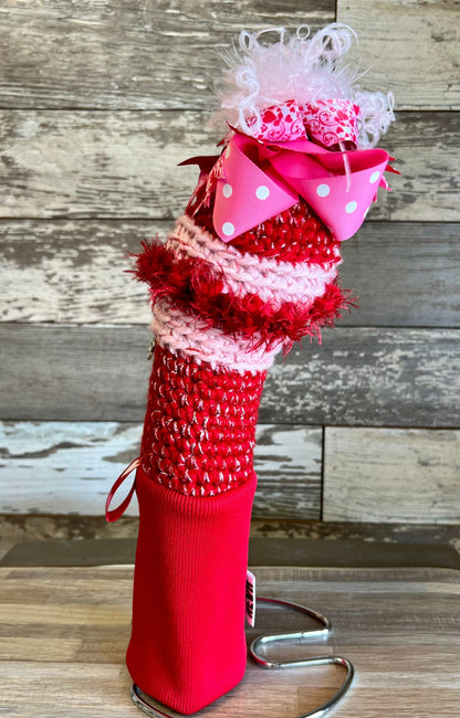 Knitted head cover titled Dream Lover featuring a red sock with a bow, jewelry, pink bow with hearts, and a red label. Handcrafted and one of a kind.